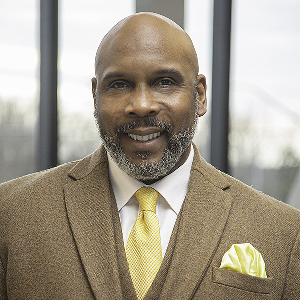 Photo of Omar Eaton-Martínez wearing a brown suit and a white shirt paired with a yellow tie with pocket square. He is bald, dark skinned, and salt and pepper bearded. He's smiling broadly and looking at the camera. 